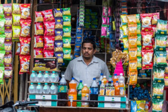किराना स्टोर में ग्राहक खरीदारी करते हुए, विभिन्न घरेलू वस्तुएं शेल्फ पर सजी हुई